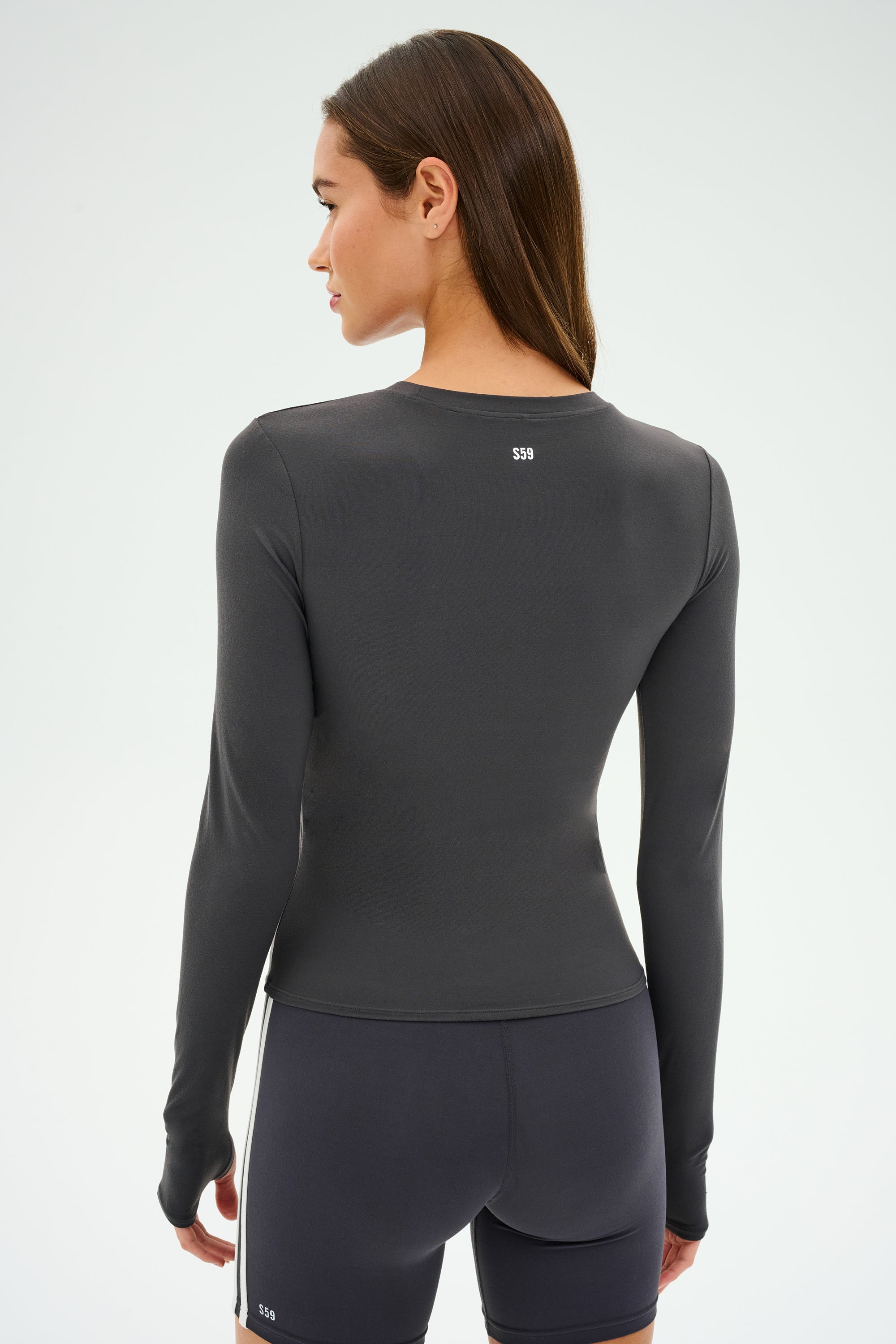 Dressed in an SPLITS59 Airweight Lite LS Waist Length top - Graphite - and shorts, a person stands facing away, ready for workouts.