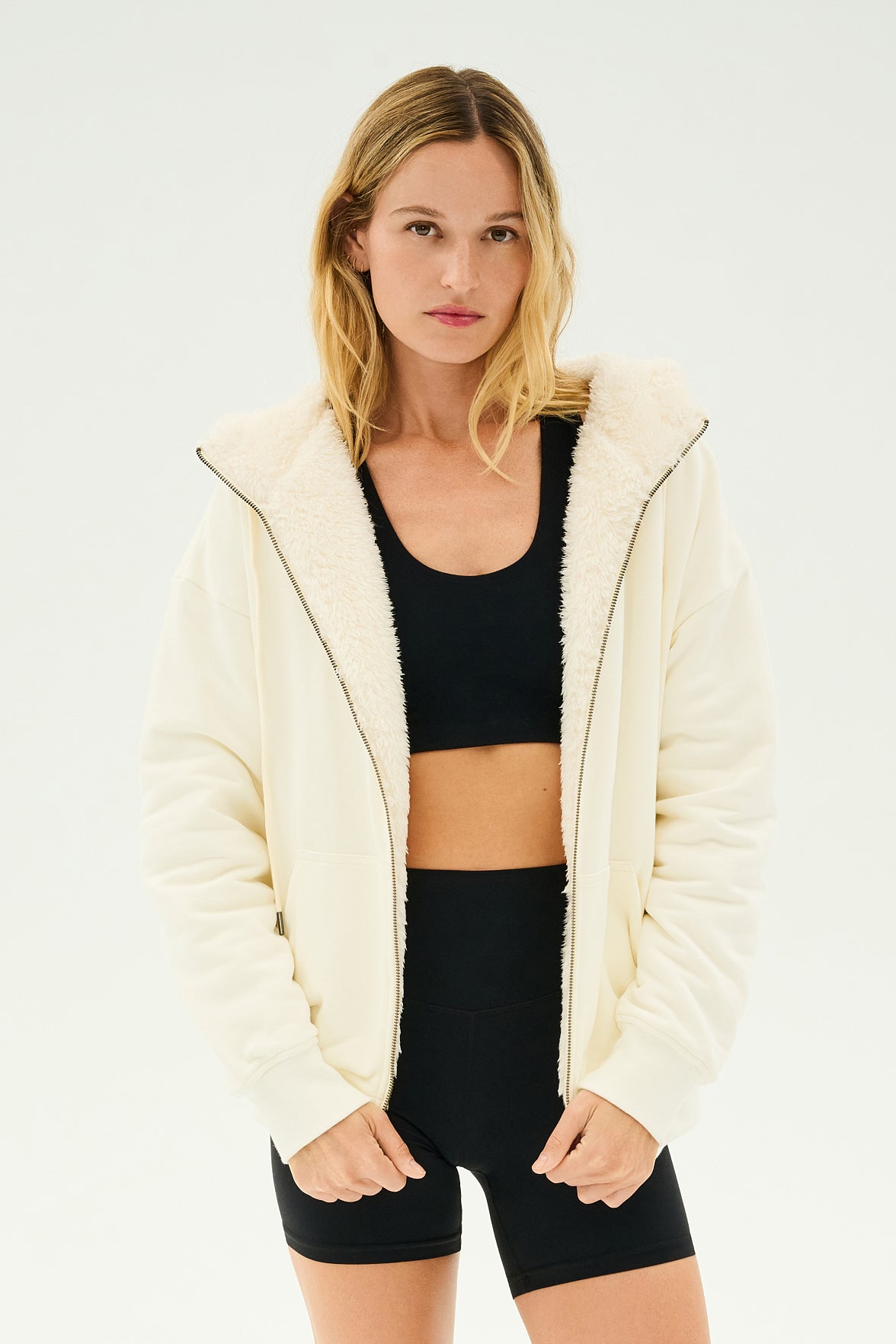 A person with long hair wears a black sports bra and shorts, layered with the SPLITS59 Lynx French Terry Sherpa Hoodie in Creme/Creme, capturing the chic comfort you'd expect après-ski. MADE IN LOS ANGELES.