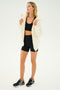 A person stands against a plain background wearing a black sports bra, black shorts, black sneakers, and the effortlessly chic SPLITS59 Lynx French Terry Sherpa Hoodie in Creme/Creme. Made in Los Angeles, this look transitions seamlessly from gym sessions to après-ski lounges.
