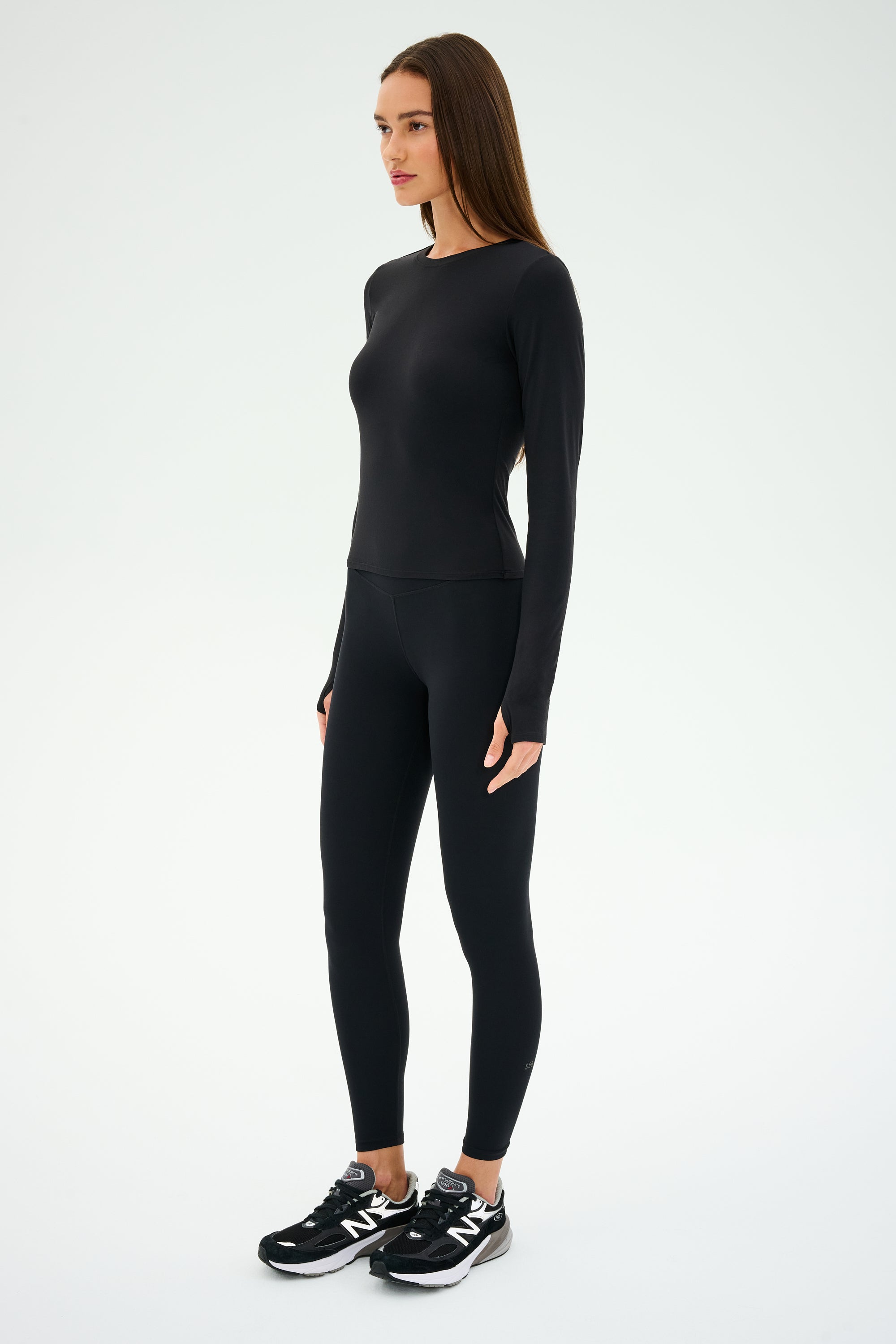 A person stands in profile, dressed for workouts in a SPLITS59 Airweight Lite LS Waist Length top, black leggings, and quick-dry black sneakers with white soles against a plain white background.