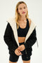 A person wearing a SPLITS59 Lynx French Terry Sherpa Hoodie in Black/Creme and a black sports outfit stands against a light background, prepared for winter workouts or ski trips.