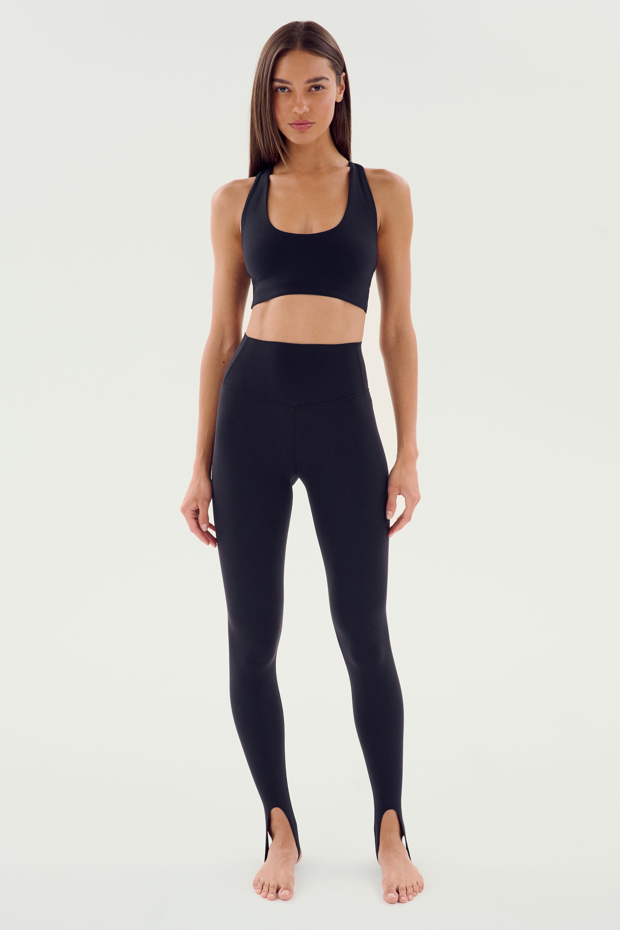 A woman stands facing forward, wearing the SPLITS59 River High Waist Airweight Stirrup leggings in black and a matching sports bra. She has long hair and a focused expression, appearing barefoot against a plain background.