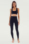 A woman stands facing forward, wearing the SPLITS59 River High Waist Airweight Stirrup leggings in black and a matching sports bra. She has long hair and a focused expression, appearing barefoot against a plain background.