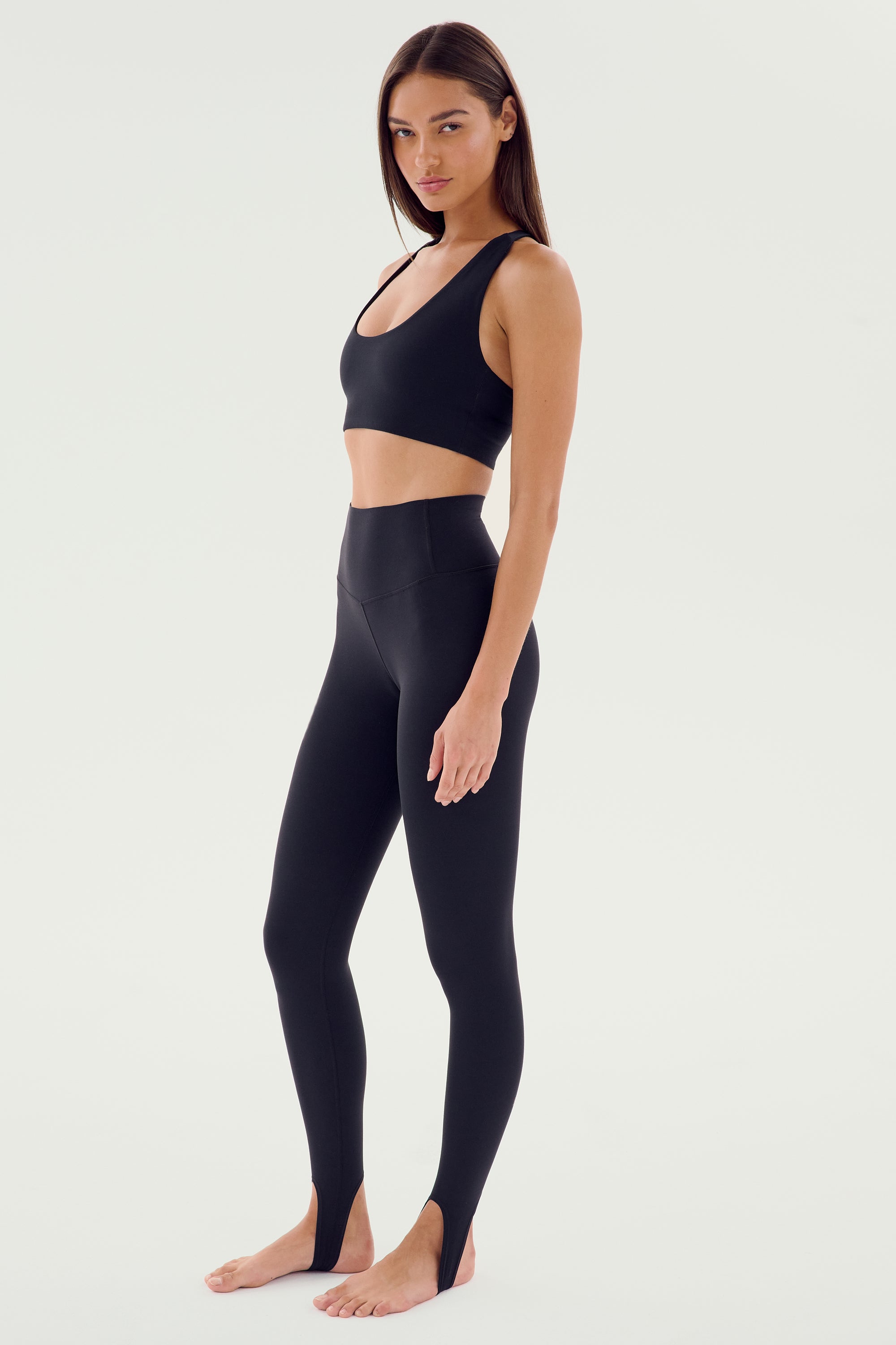 A woman is standing barefoot, wearing a black sports bra and SPLITS59's River High Waist Airweight Stirrup leggings in black, looking directly at the camera.