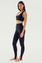 A woman is standing barefoot, wearing a black sports bra and SPLITS59's River High Waist Airweight Stirrup leggings in black, looking directly at the camera.