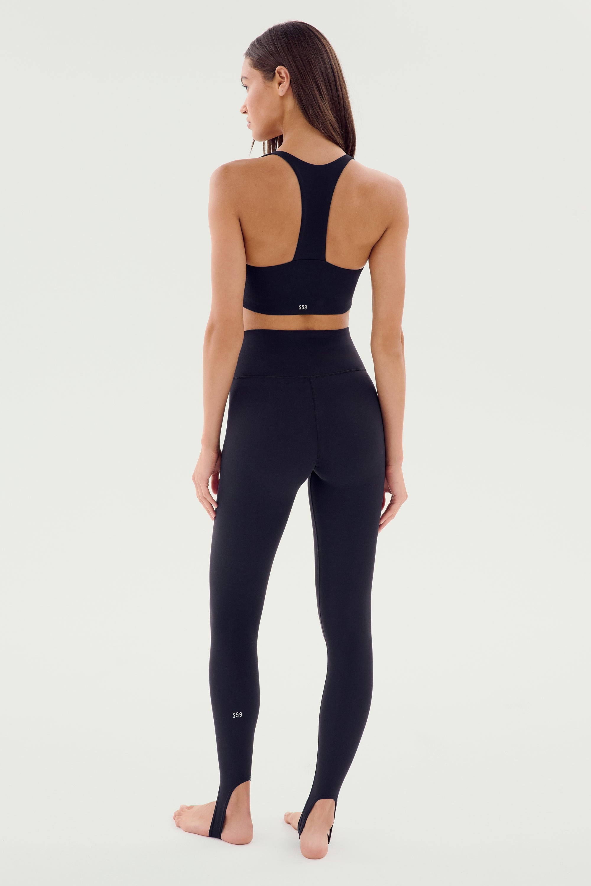 A woman in the River High Waist Airweight Stirrup leggings from SPLITS59 stands barefoot and faces away from the camera against a plain white background.