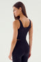 A person with long, dark hair is seen from the back wearing the black Sprint Rigor Bralette by SPLITS59 and black leggings. They are standing against a plain, light-colored background, showcasing their multi-sport performance outfit.