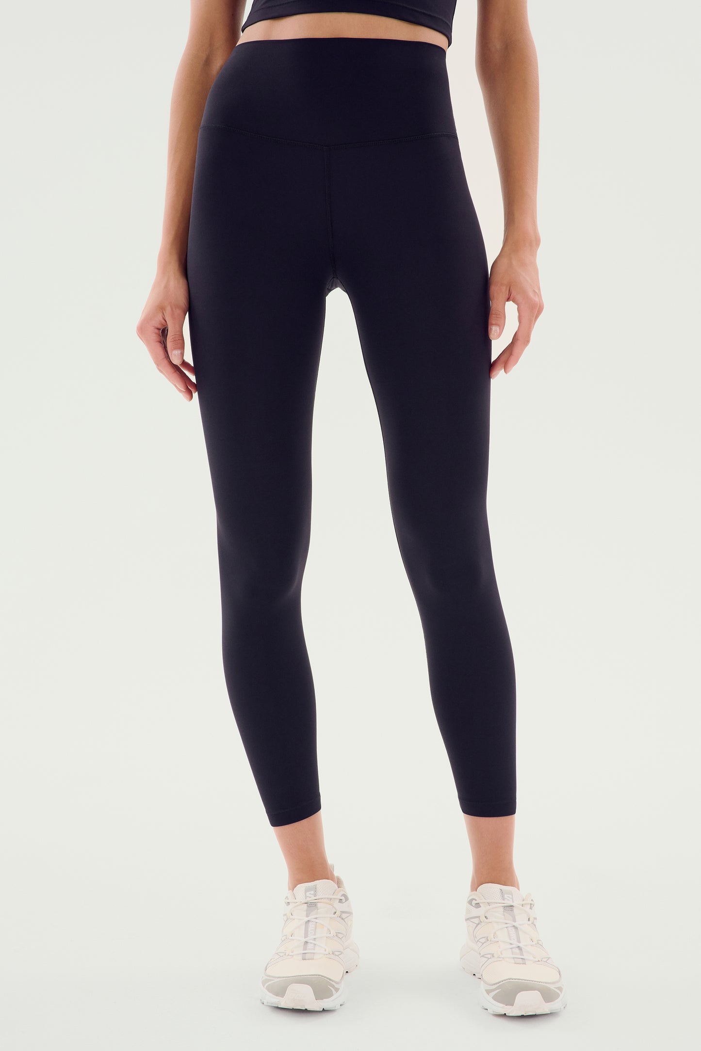 Person wearing SPLITS59 Sprint High Waist Rigor 7/8 leggings in black and white athletic shoes, standing against a plain background. Upper body partially visible.