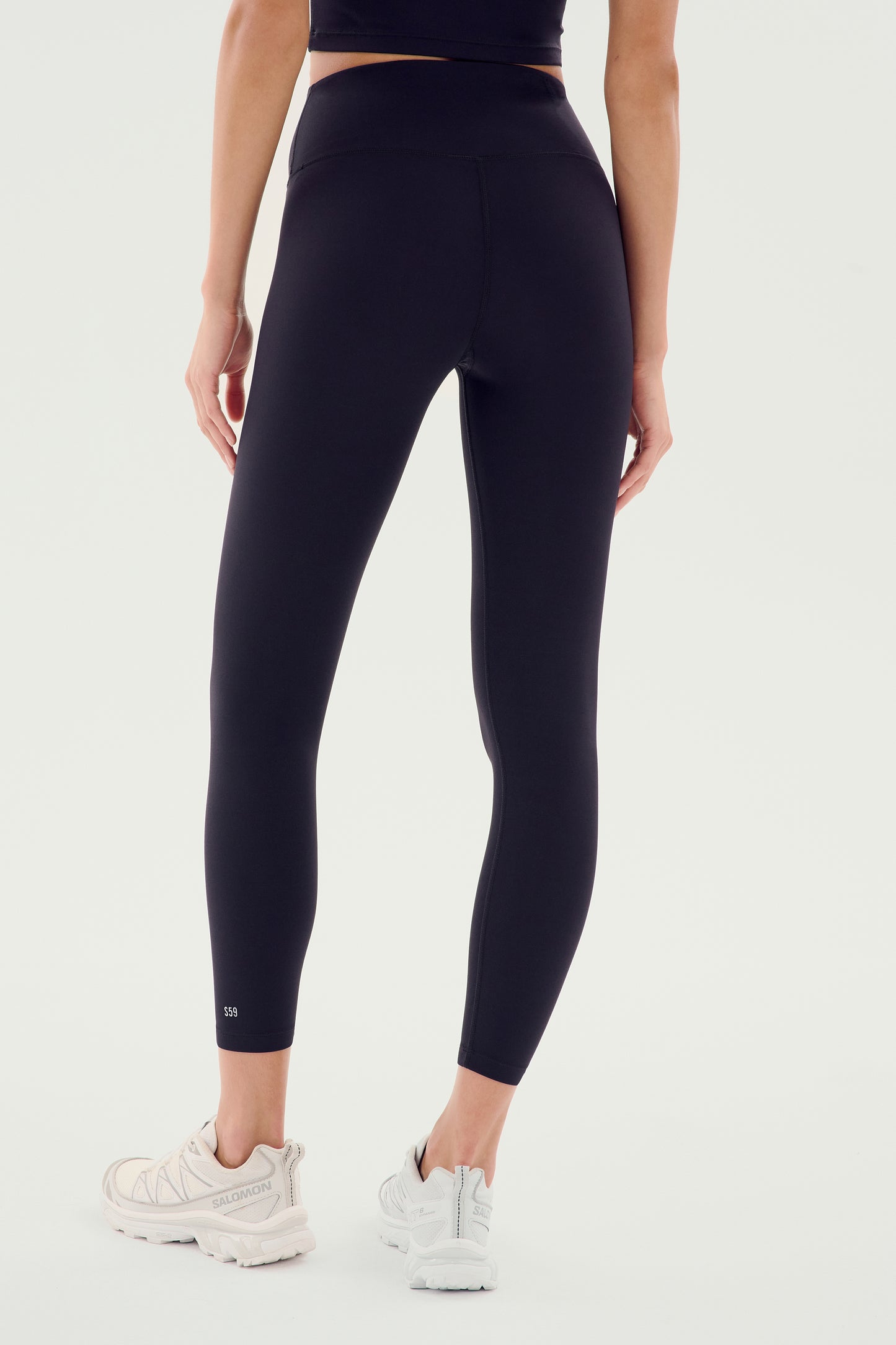 A person wearing the Sprint High Waist Rigor 7/8 leggings in black by SPLITS59 and white sneakers is standing facing away.