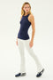 A person stands wearing a Kiki Rib Tank Full Length in Indigo from SPLITS59, white flared pants, and black sneakers against a plain white background, ready for running.
