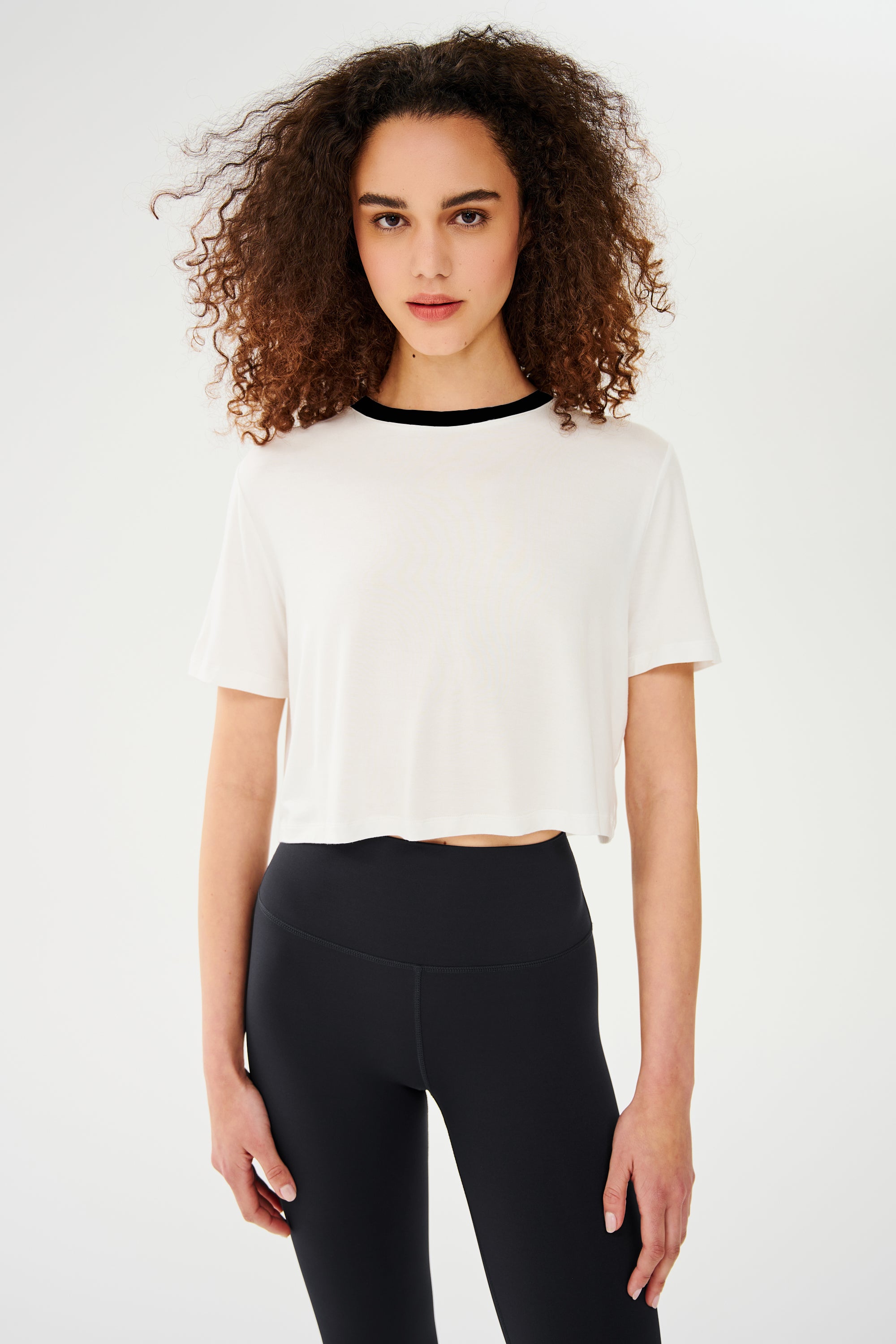 A person with curly hair wearing a white cropped Djuna Crop Ringer - White/Black by Splits59 and black leggings stands against a plain background, ready for some gym workouts.