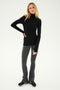A person with long hair stands wearing a black turtleneck top, Raquel High Waist Flared Legging in Heather Grey/White/Black from SPLITS59, and black shoes, with one hand on their hip.