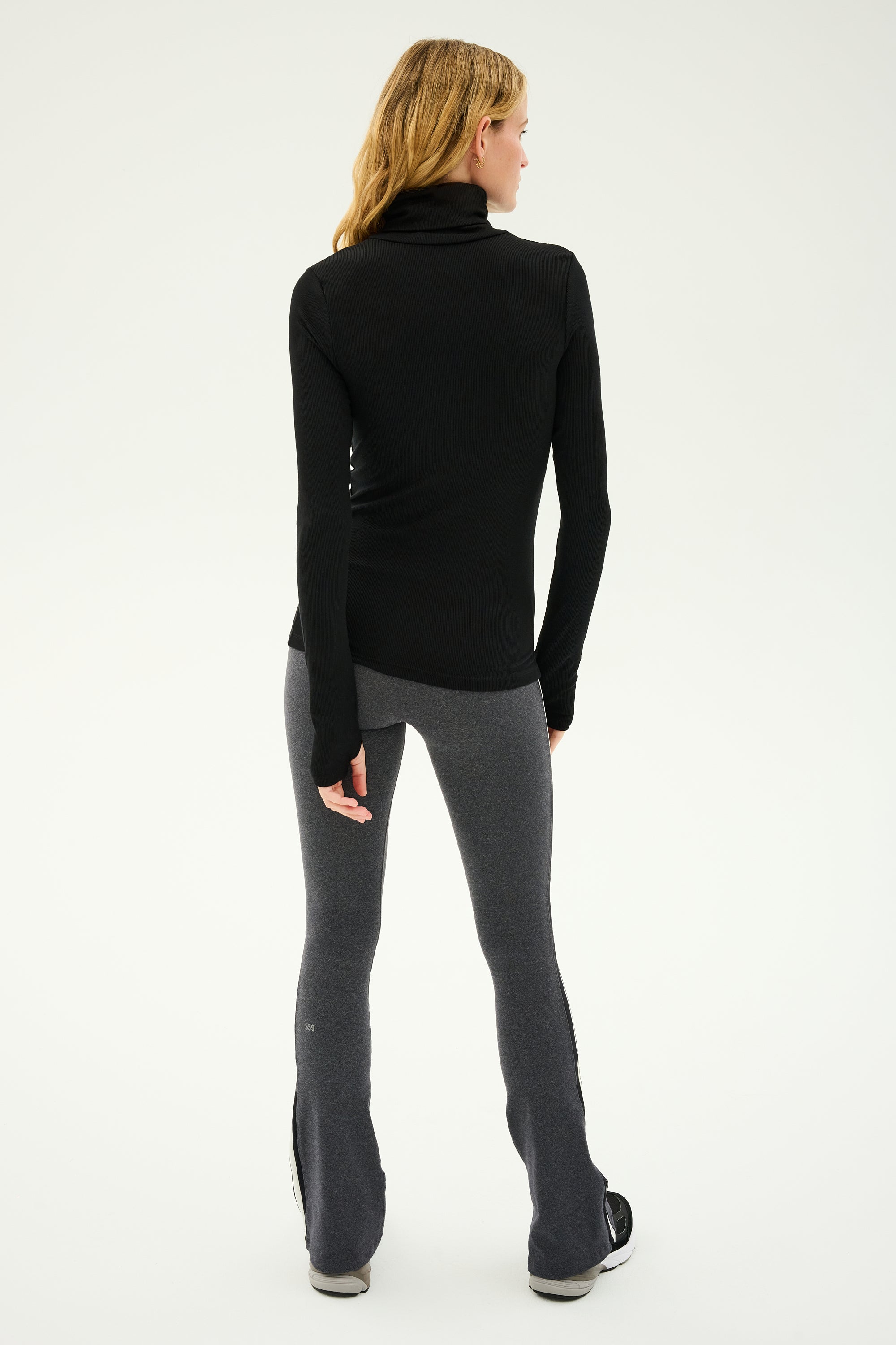 A person with long hair stands facing away, wearing a black long-sleeve turtleneck and the Raquel High Waist Flared Legging in Heather Grey/White/Black from SPLITS59, paired with black shoes.