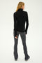 A person with long hair stands facing away, wearing a black long-sleeve turtleneck and the Raquel High Waist Flared Legging in Heather Grey/White/Black from SPLITS59, paired with black shoes.