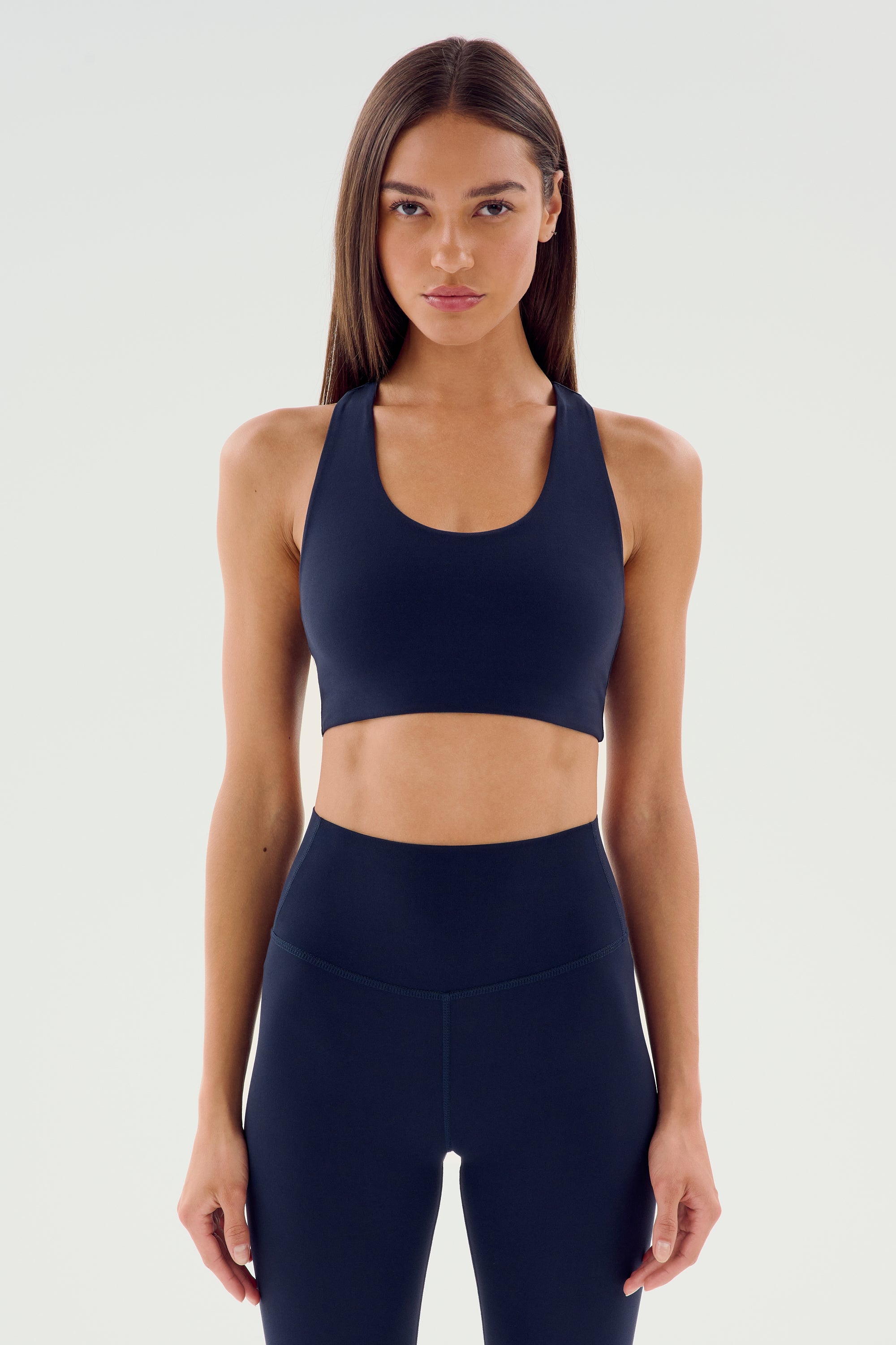 A person with long hair, dressed in an indigo Sara Airweight Bra by SPLITS59 and black leggings, stands poised against a plain light background, ready for a session of hot yoga or Pilates.