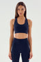 A person with long hair, dressed in an indigo Sara Airweight Bra by SPLITS59 and black leggings, stands poised against a plain light background, ready for a session of hot yoga or Pilates.