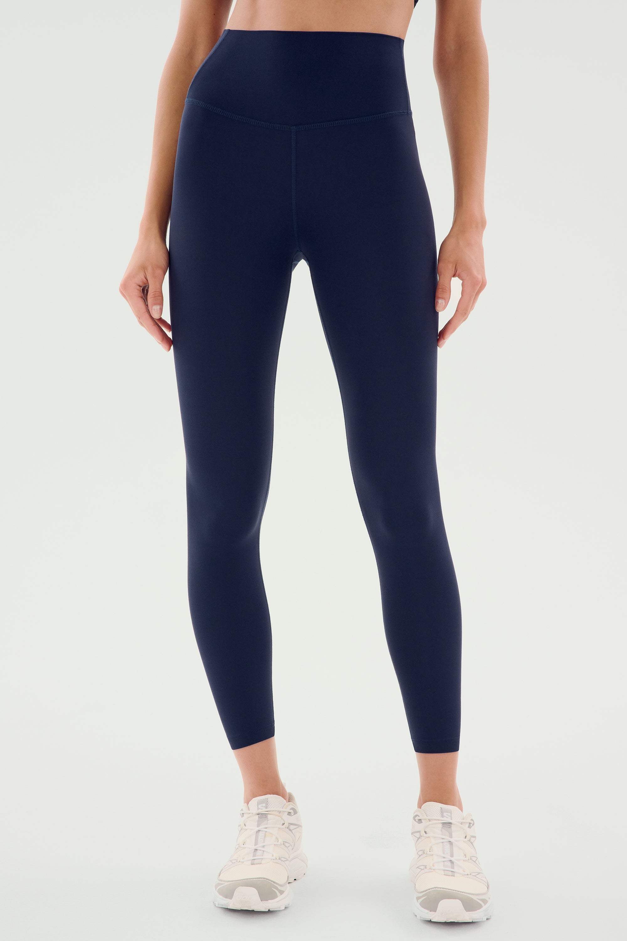 A person wearing SPLITS59's Airweight High Waist Legging in Indigo and white athletic sneakers stands against a plain background. The upper body is partially visible, with hands resting by the sides, ready for a hot yoga session.
