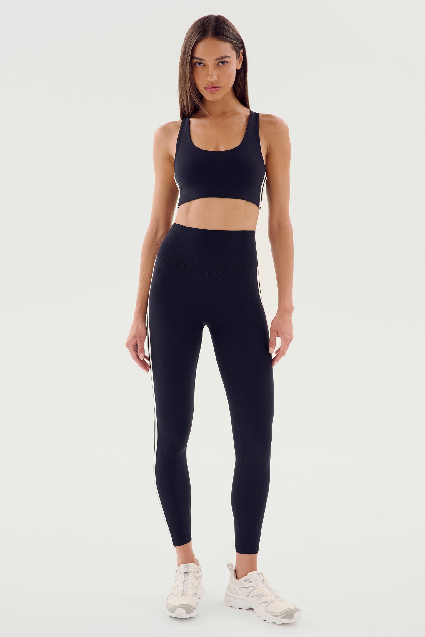 A woman faces forward in the SPLITS59 Ella High Waist Airweight 7/8 in Black/White, paired with a matching sports bra and white sneakers against a plain light background—ideal for hot yoga or everyday wear.