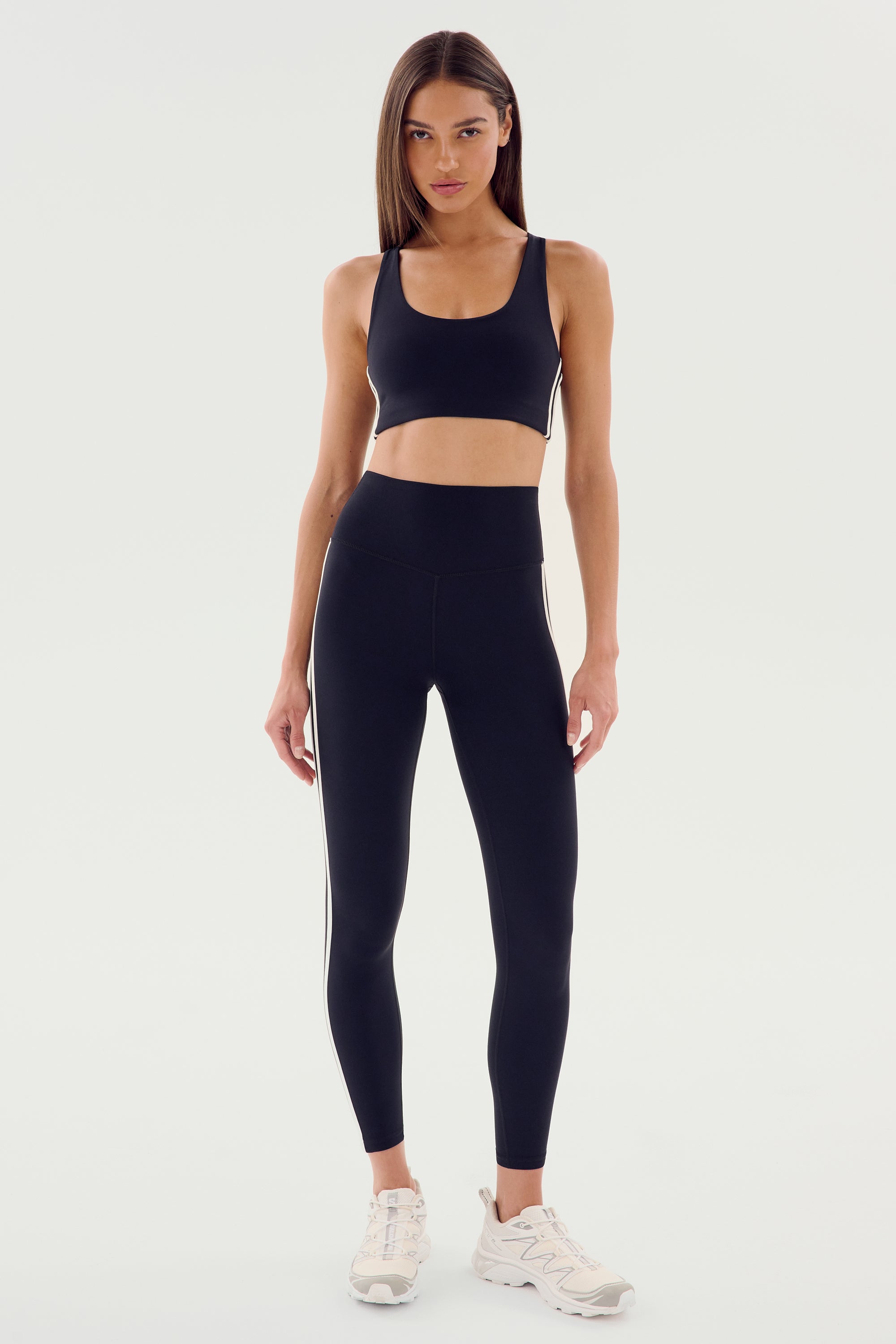 A woman faces forward in the SPLITS59 Ella High Waist Airweight 7/8 in Black/White, paired with a matching sports bra and white sneakers against a plain light background—ideal for hot yoga or everyday wear.