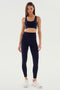 A woman faces forward in the SPLITS59 Ella High Waist Airweight 7/8 in Black/White, paired with a matching sports bra and white sneakers against a plain light background—ideal for hot yoga or everyday wear.