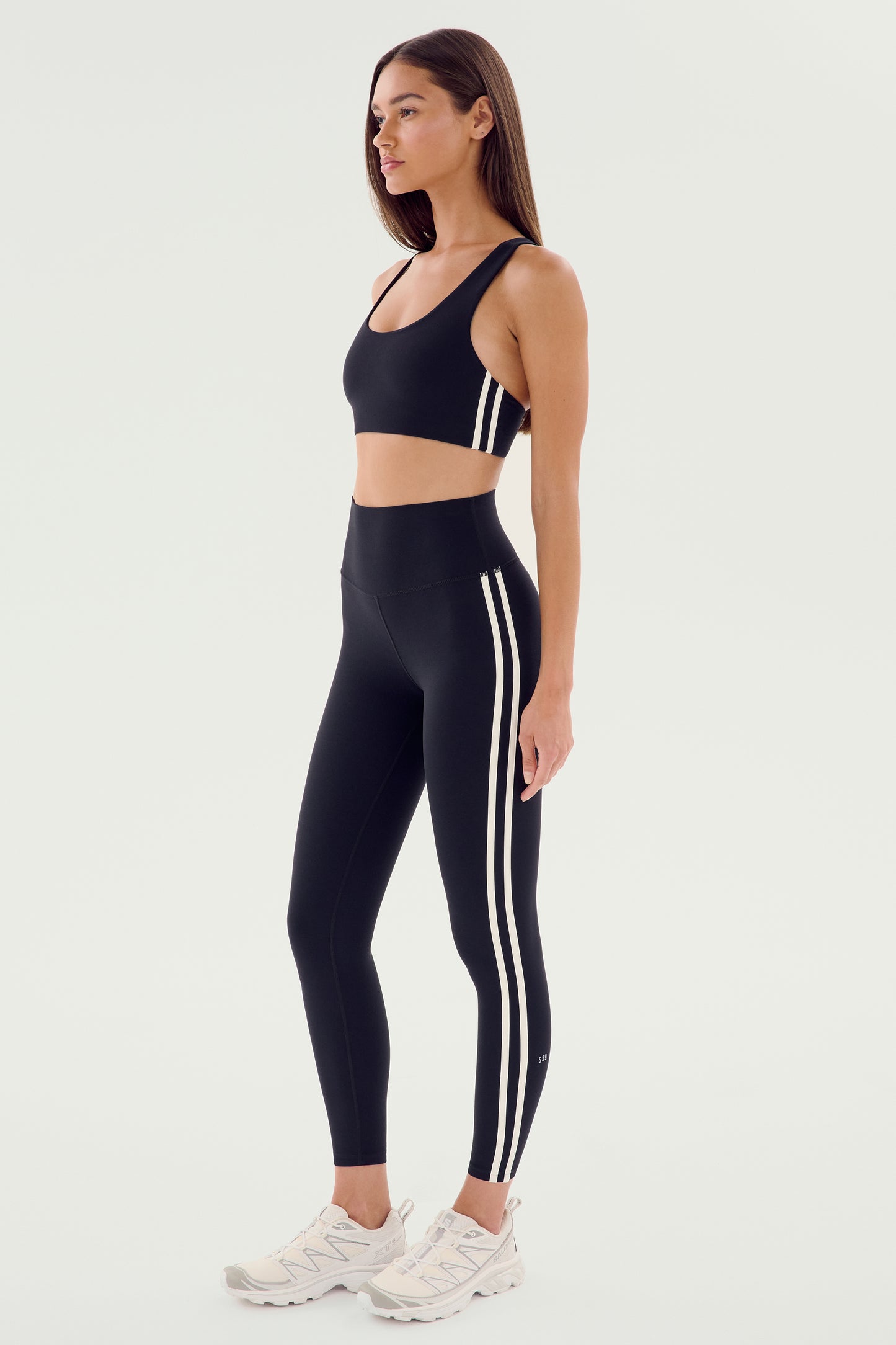A woman stands in profile wearing the SPLITS59 Ella High Waist Airweight 7/8 leggings in Black/White with matching sports bra and white sneakers—ideal for hot yoga—set against a plain background.