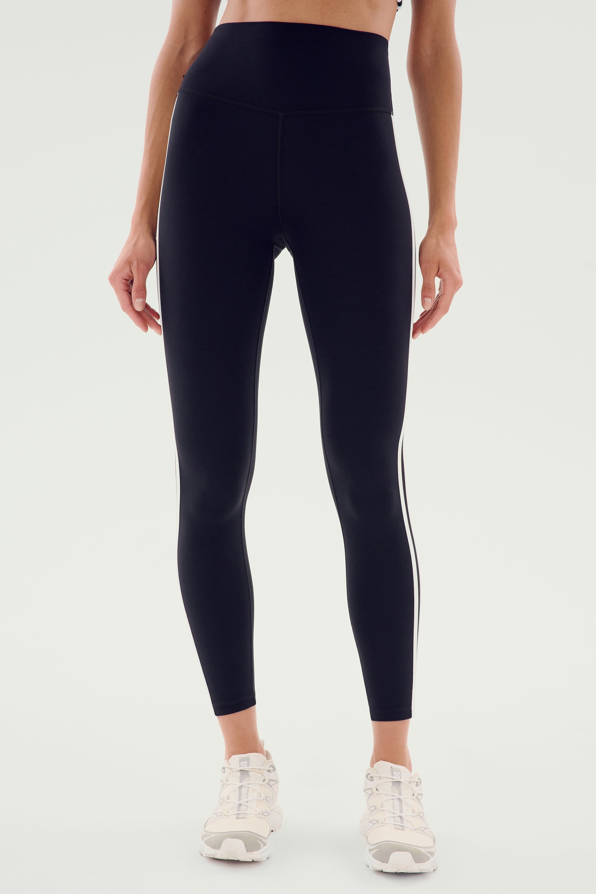 Person wearing the SPLITS59 Ella High Waist Airweight 7/8 leggings in black with white stripes on the sides, along with white sneakers, standing against a plain background.