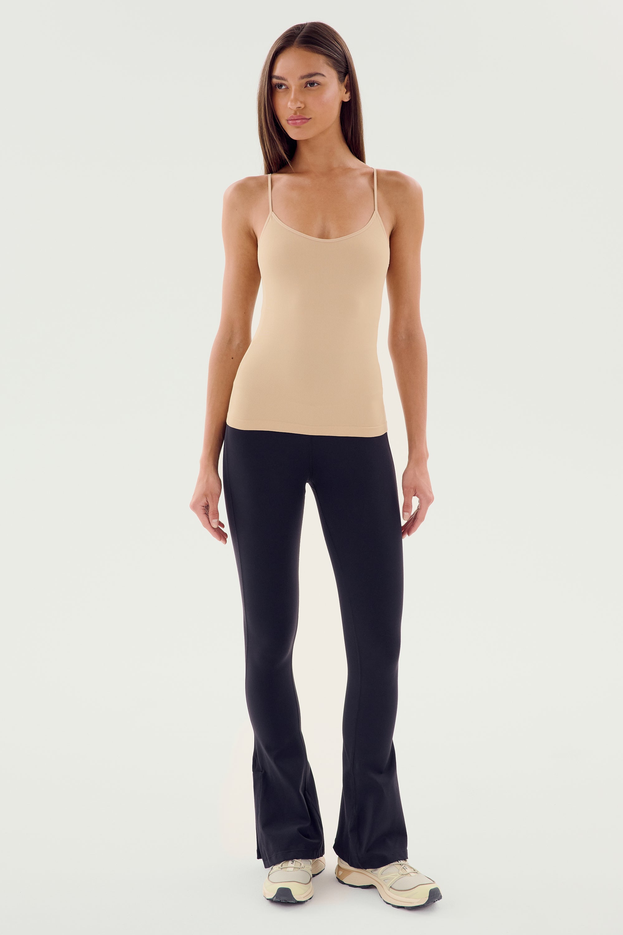 A woman stands facing forward wearing a beige tank top, SPLITS59's Raquel High Waist Flare with a stylish side split hem in black, and light-colored sneakers against a plain white background.
