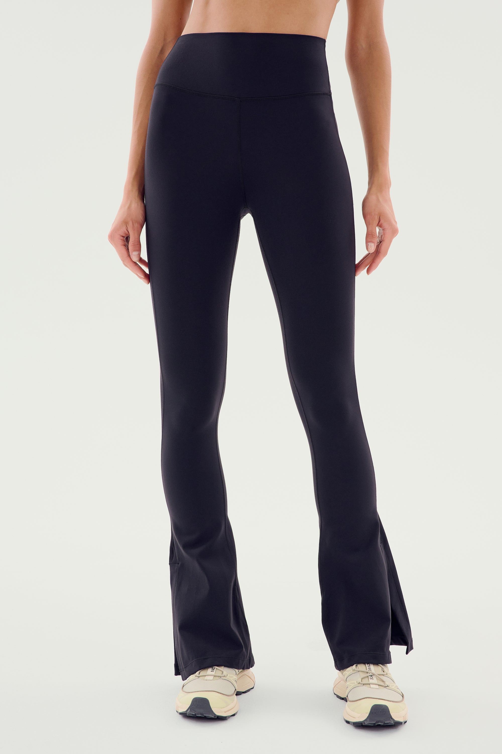 Person wearing the SPLITS59 Raquel High Waist Flare w/ Split Hem in black, paired with beige sneakers, standing against a plain white background. The stylish side split hem adds a touch of elegance to the 4-way stretch supplex fabric. Only the lower half of the body is visible.