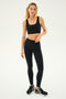 Woman wearing a black Sprint Rigor Bra by SPLITS59, crafted from ultra-luxurious Rigor fabric, paired with black leggings and running shoes, standing against a plain background.