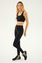 A person stands in a relaxed pose, wearing a black Sprint Rigor Bra from SPLITS59 made from ultra luxe Rigor fabric, black high-waisted leggings, and black New Balance sneakers, emphasizing their dedication to multi-sport performance against a plain white background.