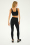 A woman is standing with her back to the camera, wearing a black Sprint Rigor Bra by SPLITS59, black leggings made from ultra-luxe Rigor fabric, and black and white sneakers.