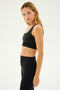 A person with long blonde hair wearing a SPLITS59 Sprint Rigor Bra in black and black leggings stands sideways facing the camera, showcasing ultra luxe Rigor fabric against a plain white background.