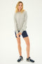 A person with long blonde hair is wearing a gray sweater, SPLITS59's Airweight High Waist Short in Indigo, and blue sneakers, standing against a plain background.