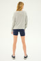A person with shoulder-length blonde hair is standing with their back to the camera, wearing a gray sweatshirt, Airweight High Waist Short in Indigo from SPLITS59, and black athletic shoes.