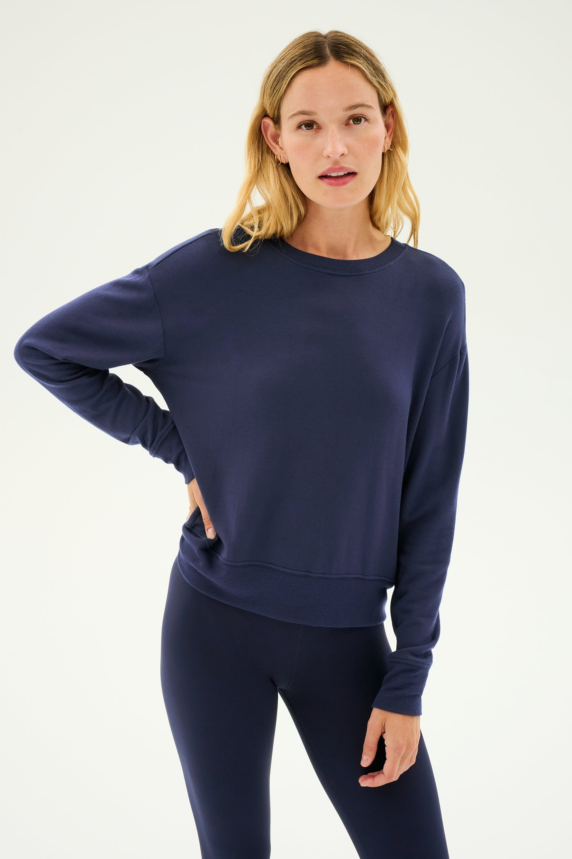 A woman stands against a plain background wearing the SPLITS59 Sonja Fleece Sweatshirt in indigo, paired with matching leggings and resting one hand on her hip.