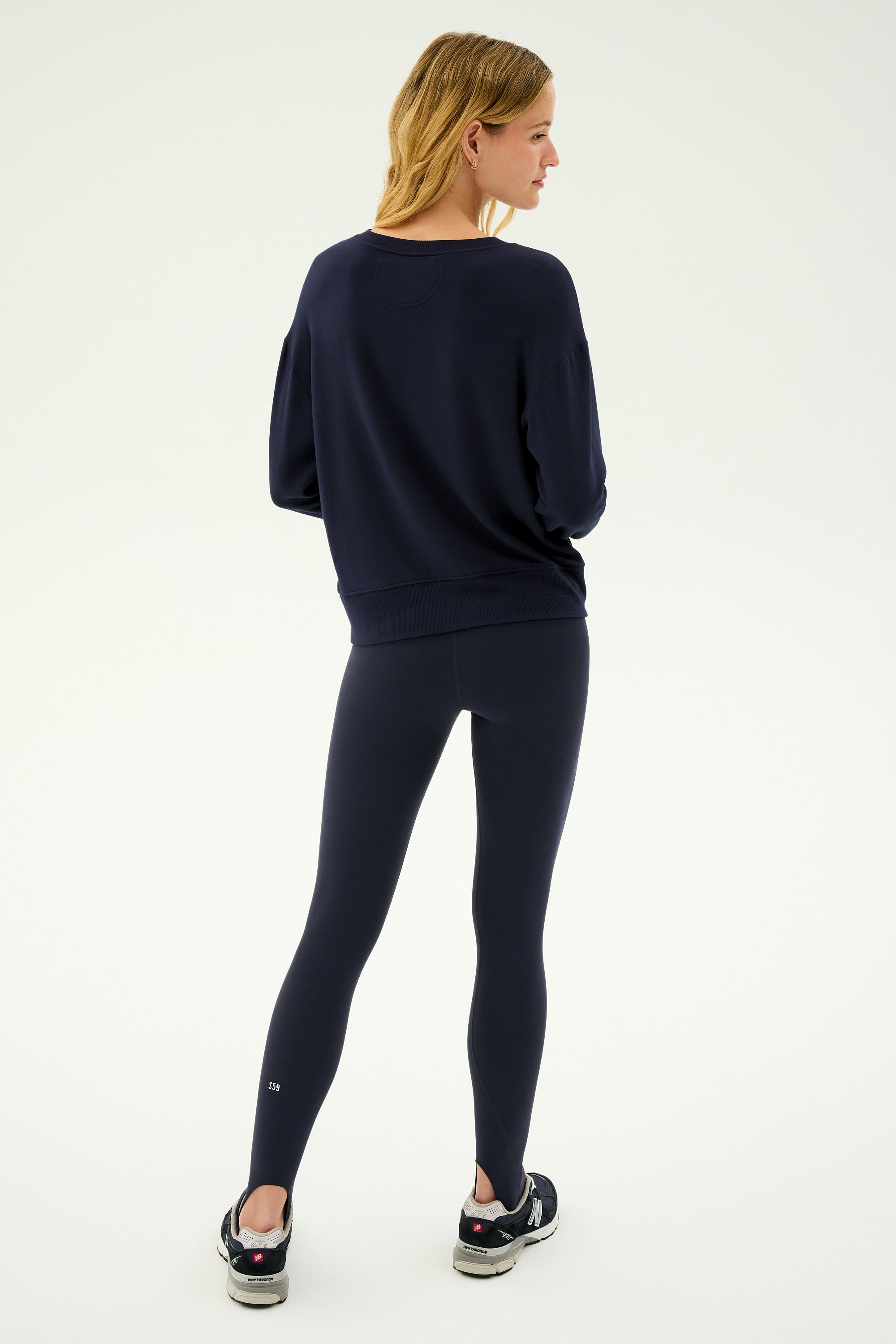 A woman stands facing away on a plain white background, wearing SPLITS59 River High Waist Airweight Stirrup leggings in Indigo, a navy sweatshirt, and black sneakers.