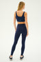 A person stands facing away, showcasing workout fitness attire in a navy blue sports bra, Sprint High Waist Rigor 7/8 leggings - Indigo by SPLITS59, and black sneakers on a light background.