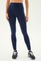 A person wearing the Sprint High Waist Rigor 7/8 leggings in Indigo from SPLITS59 and black athletic shoes stands against a plain background. Only the lower half of their body is visible, highlighting their fitness attire.