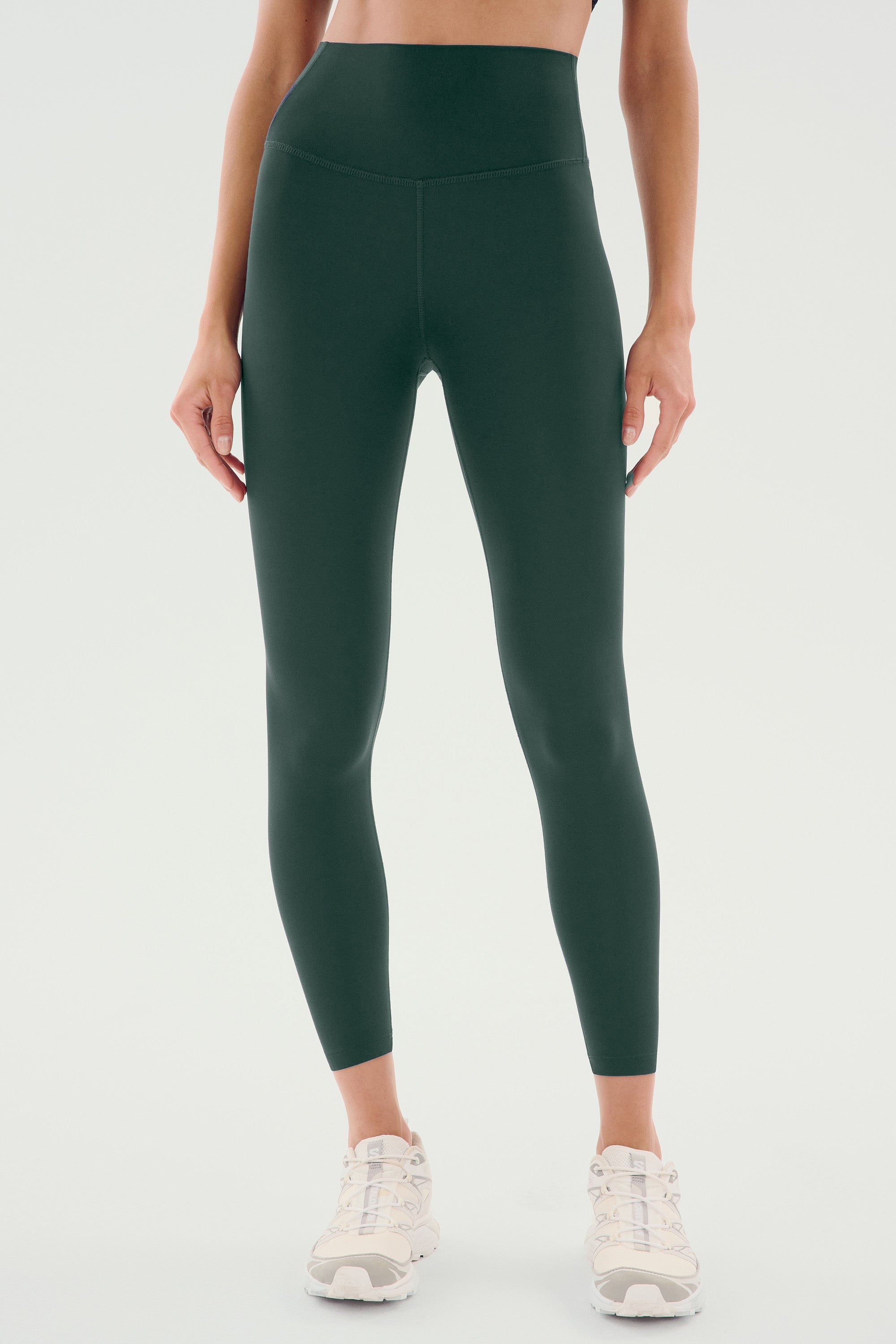 A person stands against a plain light background wearing SPLITS59 Airweight High Waist Legging - Military, crafted from ultra-soft fabric and paired with white sneakers. Ideal for hot yoga or casual wear.