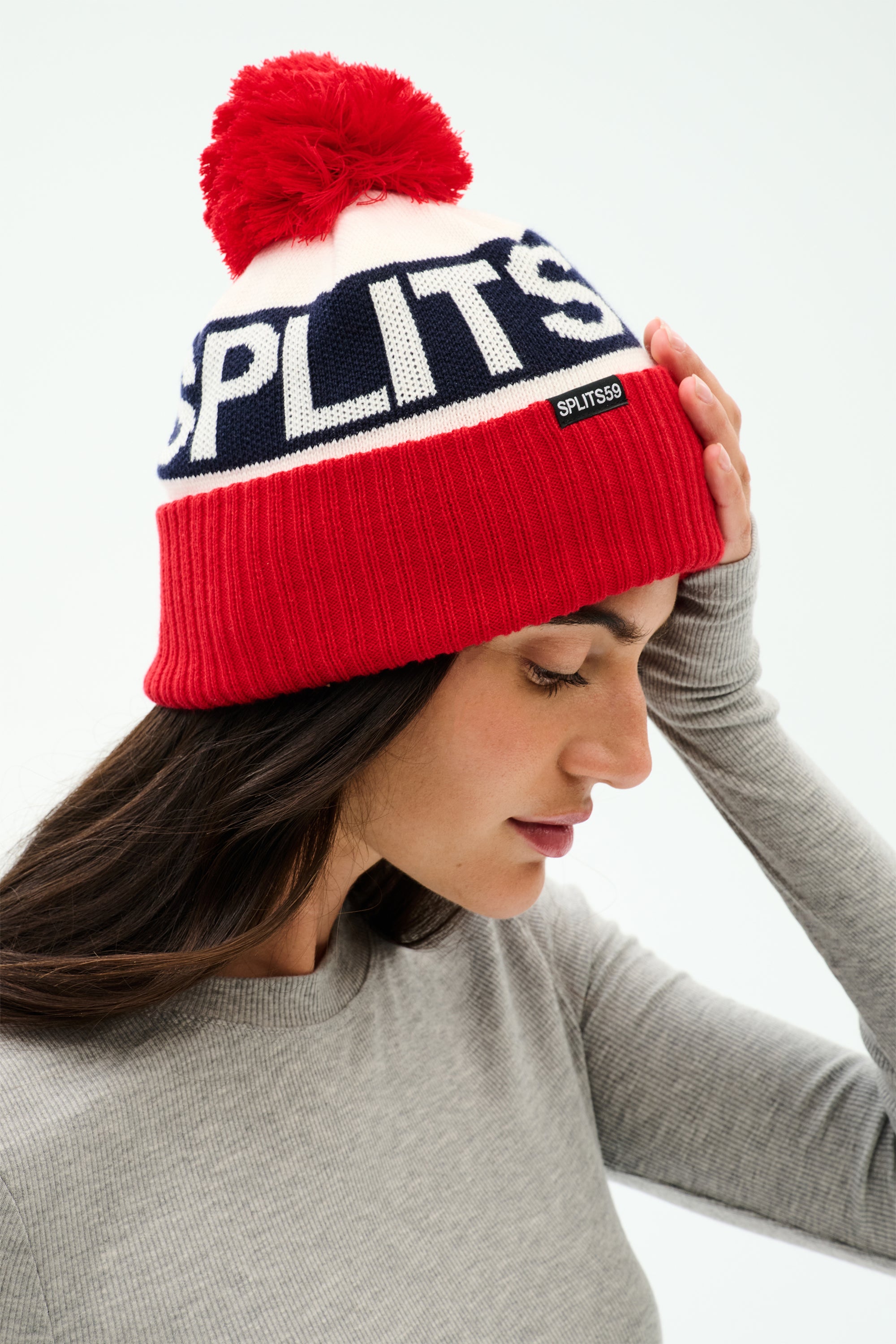 A woman in a SPLITS59 Winter Pom Pom Beanie in Lotus Red/White touches her head, ready for winter workouts in a cozy gray long-sleeve top.
