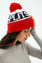 A woman wears the SPLITS59 Winter Pom Pom Beanie in red, white, and navy with logo detailing, paired with a gray ribbed long-sleeve top.