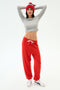 A woman stands with arms raised, wearing a SPLITS59 Winter Pom Pom Beanie in Lotus Red/White, a grey crop top, red jogger pants with white drawstring, and white sneakers—ideal winter workout attire against a plain white background.
