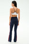 A woman faces away, wearing the SPLITS59 Loren Seamless Bra in White/Indigo, ideal for yoga and gym sessions, paired with navy flared athletic pants featuring red side stripes against a plain light background.