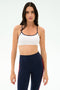 A woman stands against a plain background wearing the SPLITS59 Loren Seamless Bra in White/Indigo, perfect for gym workouts, paired with navy blue athletic leggings featuring red stripes.