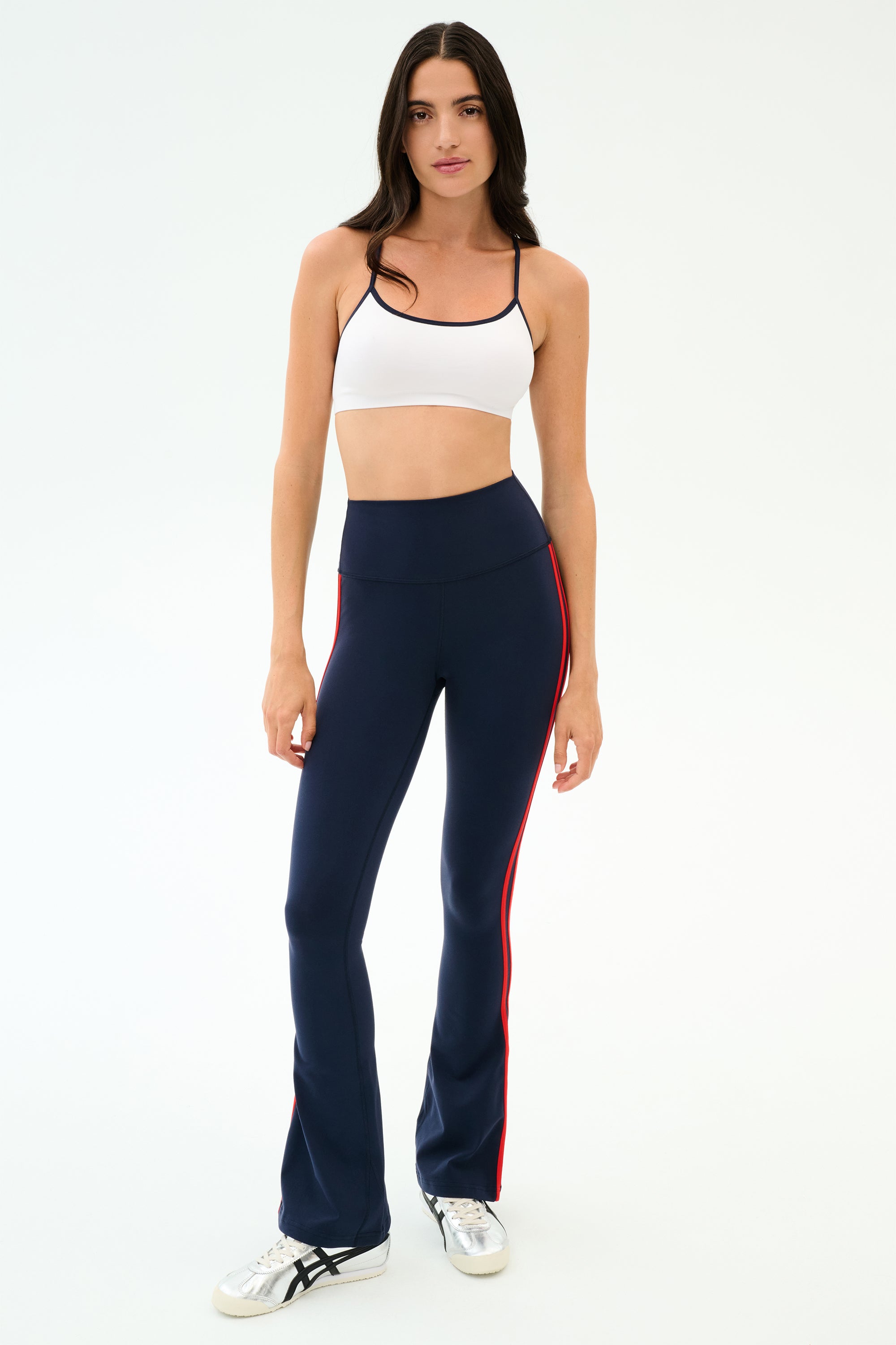A woman faces forward wearing the SPLITS59 Loren Seamless Bra in White/Indigo, navy blue flared pants with red side stripes, and white sneakers against a plain background.