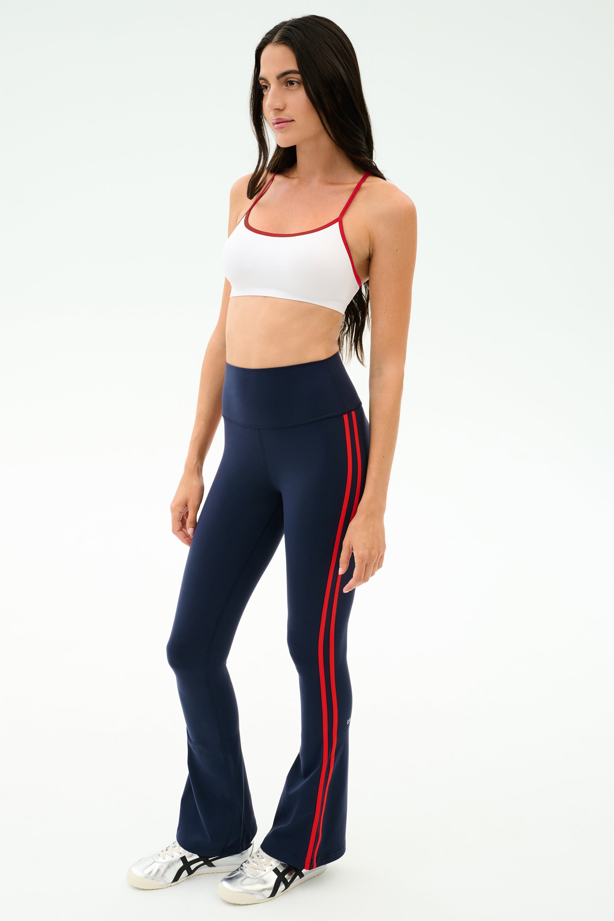 A woman wears the SPLITS59 Loren Seamless Bra in White/Lotus Red, styled with navy blue flared pants featuring red stripes and white sneakers—perfect for gym workouts or yoga—against a plain light background.