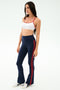 A woman wears the SPLITS59 Loren Seamless Bra in White/Lotus Red, styled with navy blue flared pants featuring red stripes and white sneakers—perfect for gym workouts or yoga—against a plain light background.