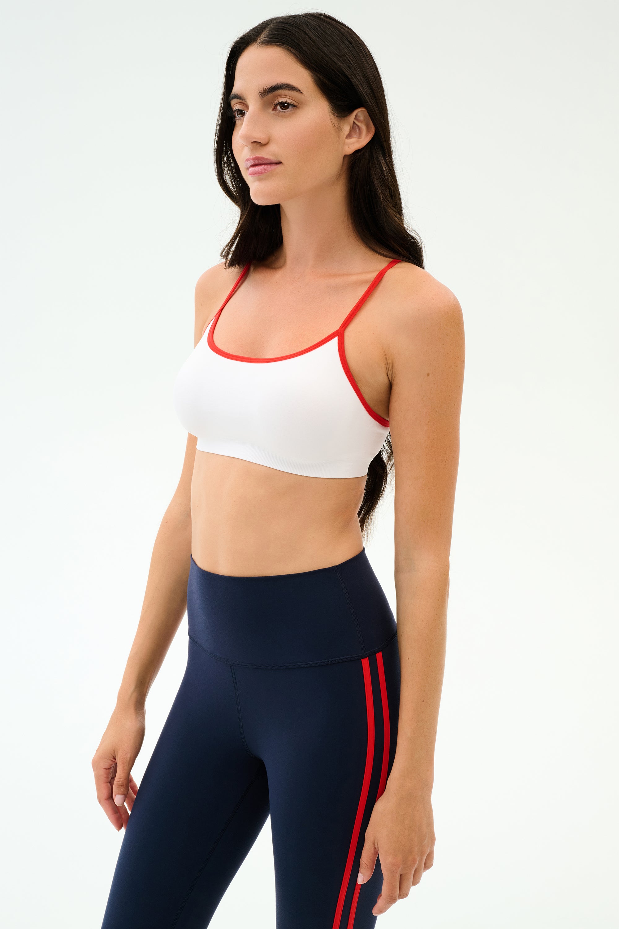 A woman wears the SPLITS59 Loren Seamless Bra in White/Lotus Red with navy high-waisted leggings featuring red stripes, perfect for gym workouts, set against a plain background.