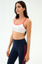 A woman wears the SPLITS59 Loren Seamless Bra in White/Lotus Red with navy high-waisted leggings featuring red stripes, perfect for gym workouts, set against a plain background.