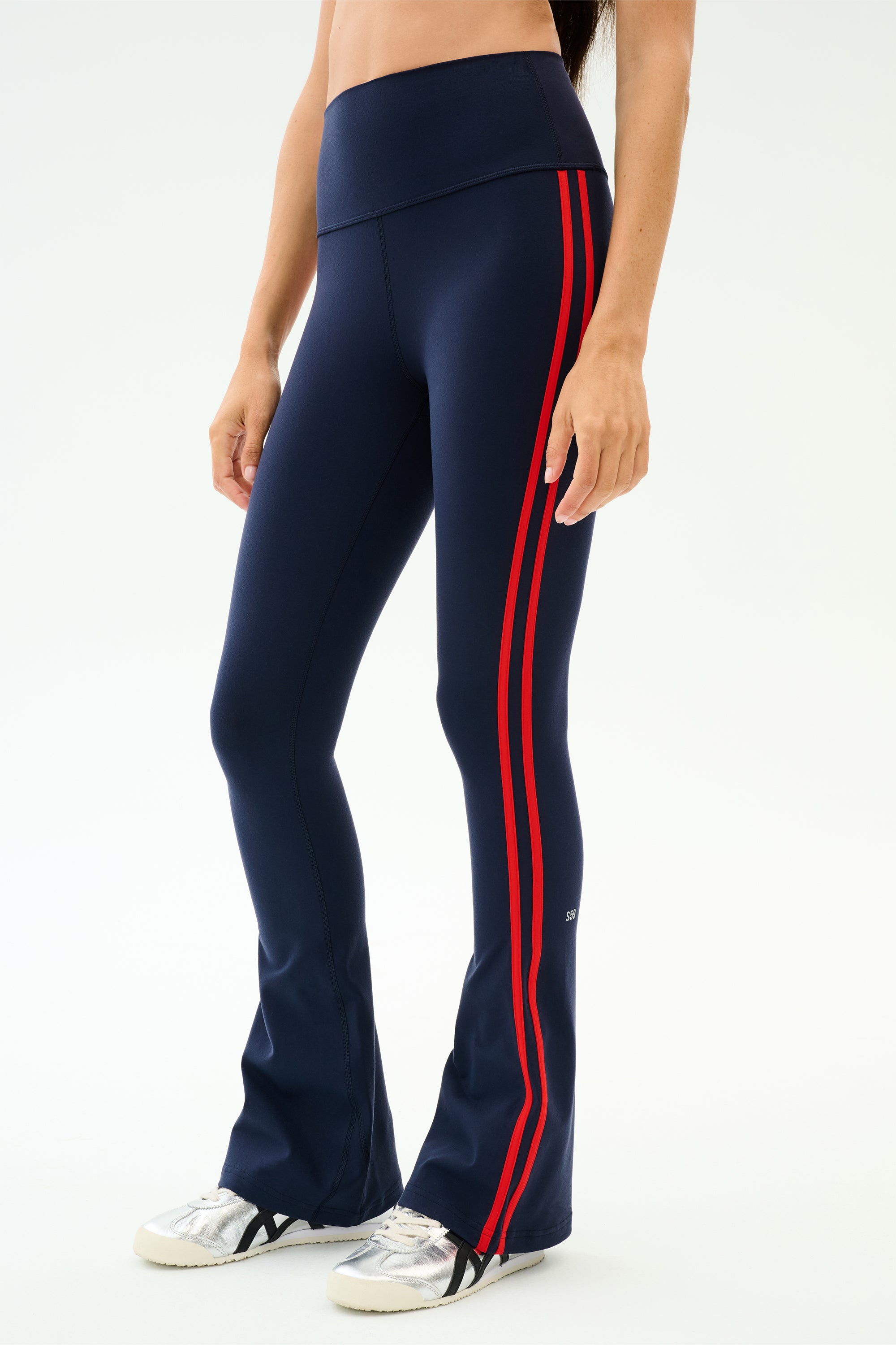 A person wears SPLITS59 Raquel High Waist Supplex Flare leggings in Indigo/Lotus Red with two red side stripes and white sneakers, standing against a plain light background. 4-way stretch fabric ensures workout versatility and comfort.